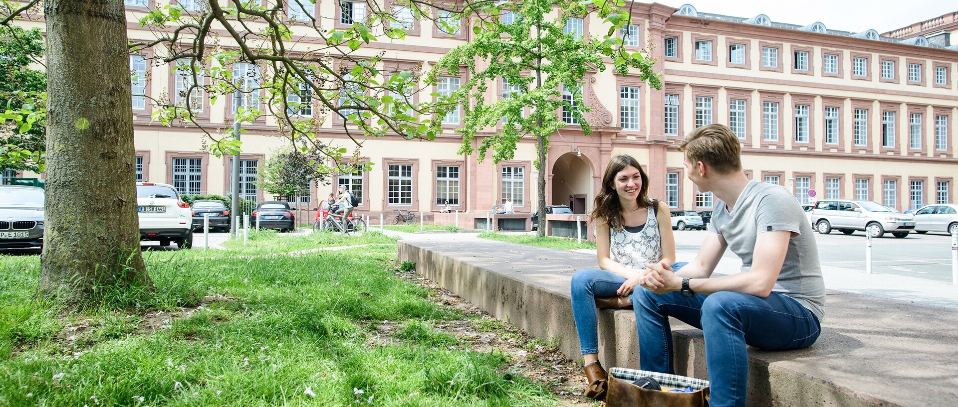 Two students sit on a stone in front of the west wing of the University of Mannheim and talk.
